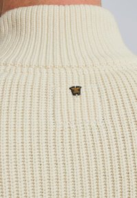 Crème-kleurige gebreide trui met een hoge kraag en ribbelstructuur, met een zwart en gouden logo op het achterpand.