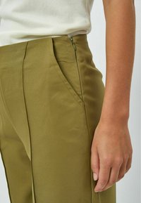 Pantalon ajusté vert olive avec une texture lisse et des poches latérales. Dispose d'une fermeture éclair discrète à la taille.