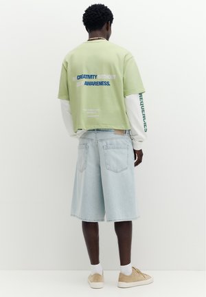 Homme porte une chemise à manches courtes vert clair par-dessus une chemise à manches longues blanche et un short en jean bleu clair, debout devant un fond blanc uni.