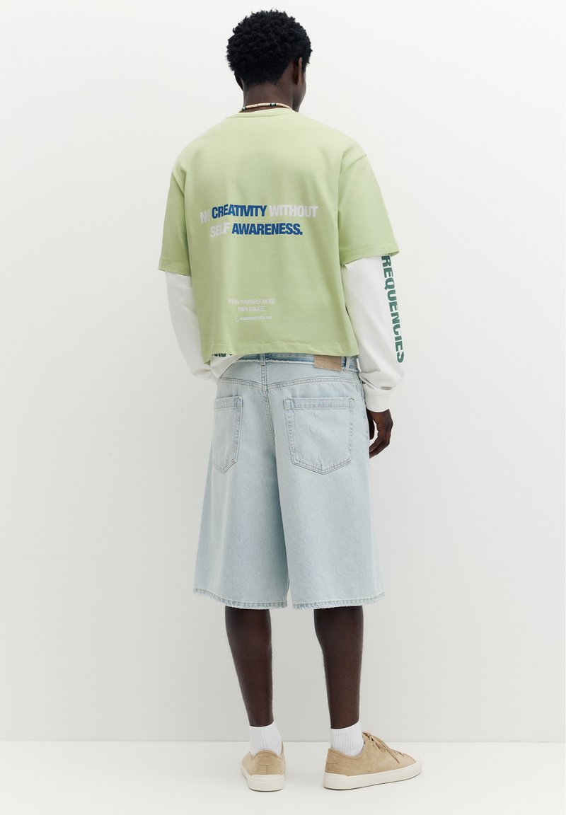 Homme porte une chemise à manches courtes vert clair par-dessus une chemise à manches longues blanche et un short en jean bleu clair, debout devant un fond blanc uni.