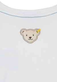 Camicia bianca in cotone con un logo di un orso ricamato e testurizzato in beige con dettagli grigio chiaro e un piccolo etichetta gialla sull'orecchio.