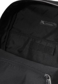 Scomparto interno dello zaino nero con tessuto liscio, etichetta Calvin Klein e una tasca con zip dotata di cinghie per il fissaggio degli oggetti.