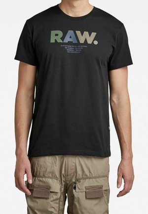 Mann trägt ein schwarzes T-Shirt mit der Aufschrift "RAW." in grünen, blauen und beigen Buchstaben sowie beige Cargohosen mit Reißverschluss- und Klappentaschen.