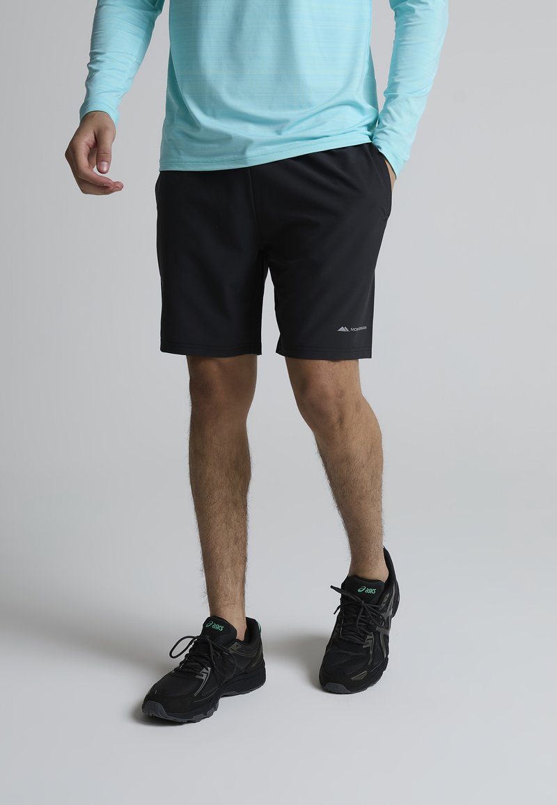 Homme portant un short de sport noir et des chaussures de course noires, debout, une main dans la poche et l'autre détendue le long du corps.
