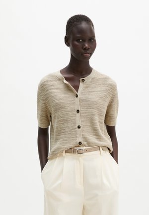 Modella indossa un cardigan beige a maniche corte in maglia con bottoni e pantaloni plissettati color avorio con cintura beige, mani nelle tasche, sfondo neutro.