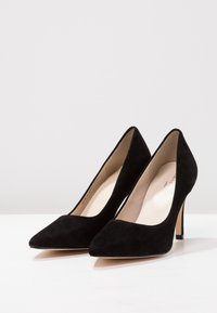 Escarpins en suede noir avec un bout pointu et un talon aiguille fin. L'intérieur présente une doublure beige lisse.