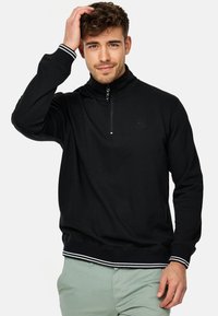Svart hettegensere med glidelås og høy krage, med hvite striper på ermene og kanten. Glatt stoff, avslappet passform.