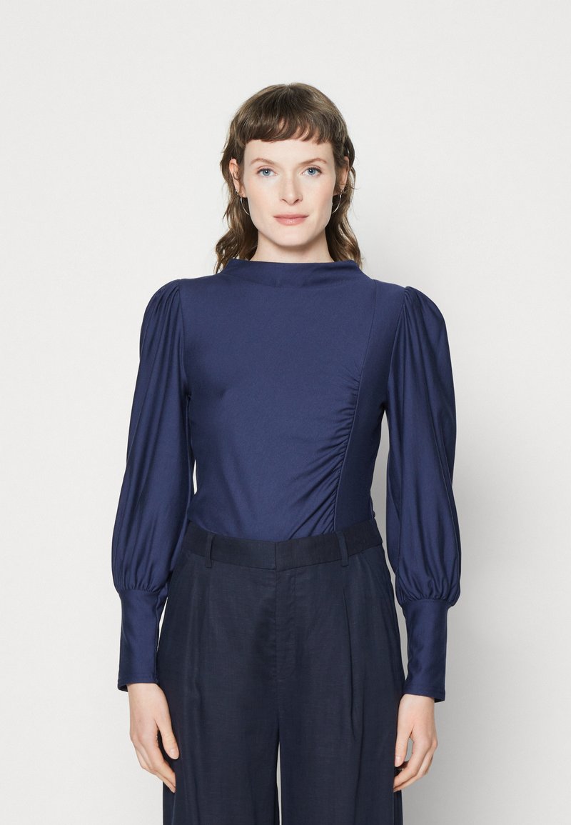 Navy Langarm-Bodysuit mit hohem Kragen, gerafften Seitendetails und puffigen Ärmeln, kombiniert mit dunklen Leinenhosen. Glatte Textur.