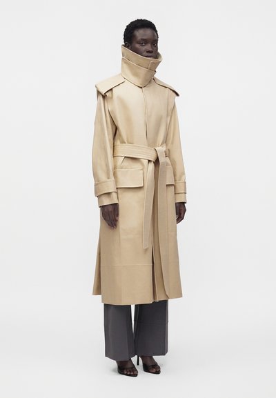 Personne à la peau foncée portant un trench-coat beige ceinturé avec un col montant et un pantalon gris à jambes larges, debout sur un fond blanc uni.