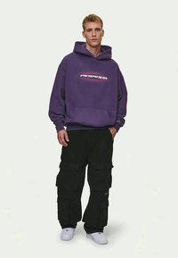 Felpa con cappuccio viola, dotata di tasca frontale e logo bianco. Abbinata a pantaloni cargo neri con più tasche e sneakers bianche.