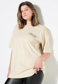 Béžové oversized tričko vyrobené z bavlny, se multikulturním textem "PŘICHÁZÍ TO VE VLNÁCH" v zakřiveném designu na levé části hrudi.