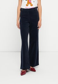 Vrouw in een hooggewaisted marineblauwe corduroy wijde broek en rode puntige veterschoenen tegen een effen witte achtergrond.