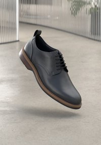 Scarpa da uomo in pelle nera con lacci e suola marrone sospesa sopra un pavimento liscio grigio chiaro, con un sfondo di corrimano in metallo sfocato.