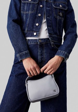 Mains tenant un petit sac à main Calvin Klein gris clair sur un jean denim bleu foncé et une veste en denim assortie avec une chemise blanche en dessous.