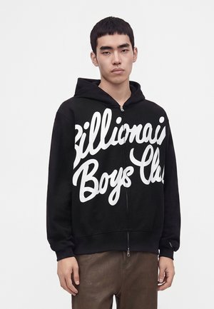 Jeune homme portant un sweat à capuche noir zippé avec un texte en écriture blanche "Billionaire Boys Club" et un pantalon marron sur un fond clair uni.