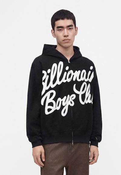 Млад мъж, носещ черна спортна блуза с цип с бял текст на скрипт "Billionaire Boys Club" и кафяв панталон на светъл фон.