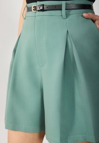 Shorts taille haute à plis en tissu turquoise clair à texture lisse, dotés d'une ceinture noire et d'une coupe classique et ajustée.