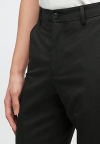 Črne po meri krojene hlače z gladko teksturo, zadaj s sprednjim žepom, zankami za pas in zaponko na gumb. Nosen s svetlo obarvano srajco.