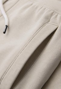 Gros plan sur le tissu beige d'un sweat-shirt avec une poche à bord-côte et une cordelette blanche à embout noir.