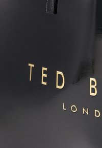 Saco tote preto brilhante com lettering dourado em relevo "TED BAKER LONDON". Apresenta uma textura suave e um design estruturado.