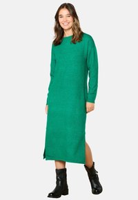 Abito verde lavorato a maglia con maniche lunghe e scollo rotondo, caratterizzato da uno spacco laterale. Abbinato a stivaletti neri alla caviglia per un contrasto.