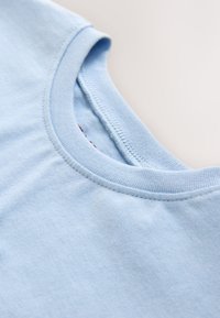 Detailní záběr modrého bavlněného trička ukazující jemné šití a hladkou strukturu látky na neutrálním pozadí.