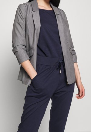 Frau trägt marineblaues Freizeit-Top und Hose mit einem hellgrauen Blazer, steht mit einer Hand in der Tasche vor einem einfarbigen Hintergrund.