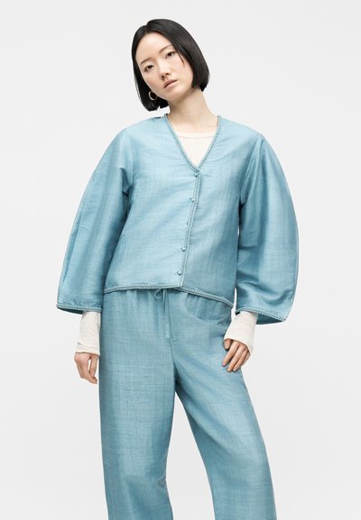 Νεαρή γυναίκα με κοντά μαύρα μαλλιά φοράει φαρδύ, ανοιχτό μπλε υφαντό σακάκι με κουμπιά και ταιριαστό παντελόνι με φαρδιά παντελόνα, με λευκά μανίκια από μέσα.