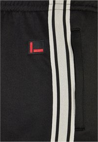 Svarte treningsshorts med teksturert stoff, en liten brodert logo i rødt og svart, og to vertikale hvite striper på siden.