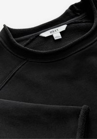 Sweat-shirt noir à col rond avec couture visible sur le col et une étiquette blanche portant la mention "Next" et "Made in Bangladesh."