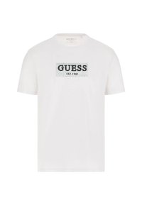 White cotton t-shirt featuring a rectangular black "GUESS" logo with "EST. 1981" underneath, set against a textured light blue background.