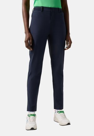 Personne portant un pantalon bleu marine ajusté, des baskets blanches à lacets verts, et un haut vert vif, debout devant un fond uni.