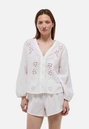 Femme portant un chemisier blanc à manches longues boutonné avec broderie florale découpée et short assorti sur fond uni.