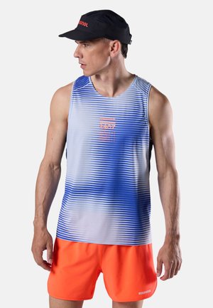 Hombre con camiseta sin mangas a rayas azules y blancas, pantalones cortos naranja brillante, gorra negra, mirando hacia la izquierda contra un fondo liso.