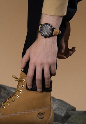 Montre noire avec fonction chronographe et bracelet en cuir tan, associée à une botte tan dotée d'un col noir et d'œillets en métal.