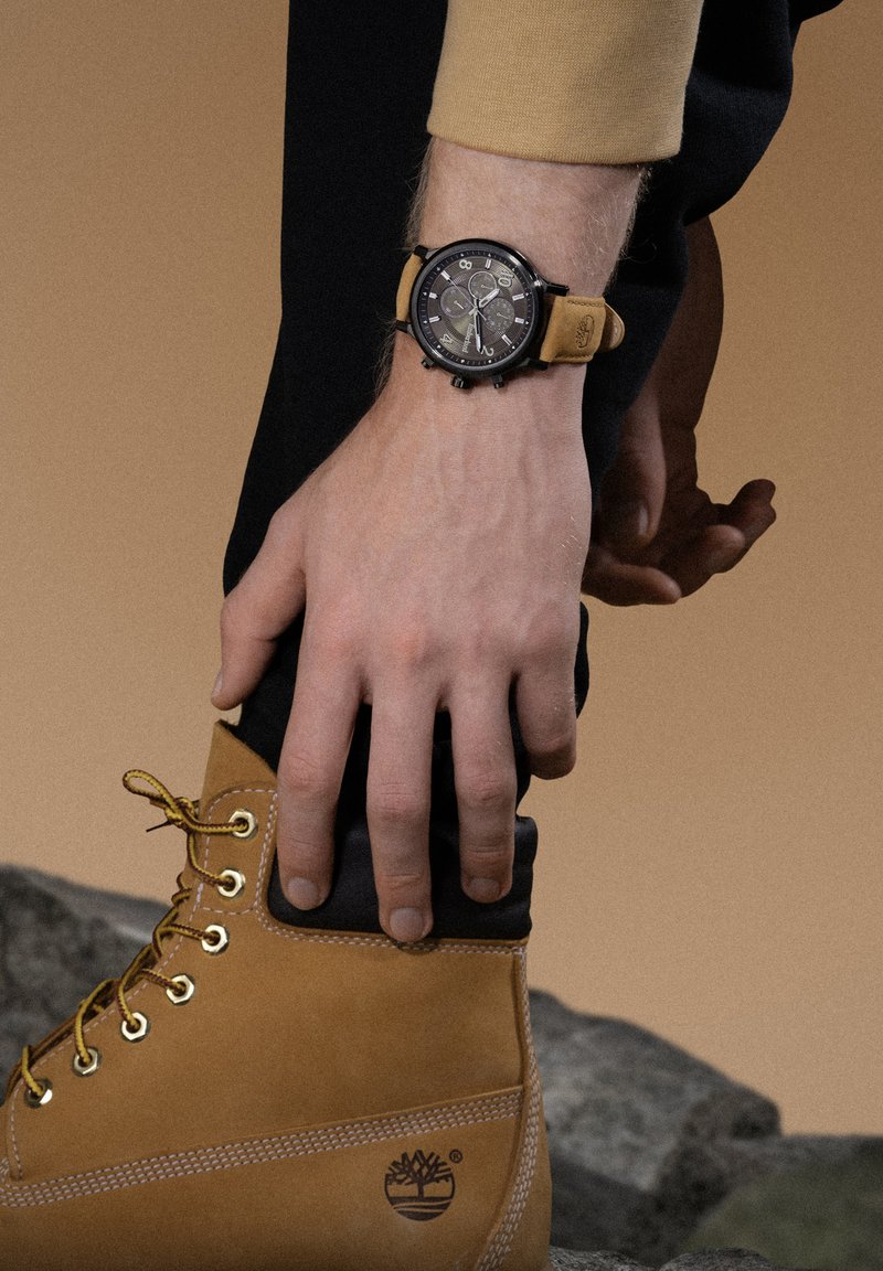 Schwarze Uhr mit Chronographenfunktion und braunem Lederarmband, kombiniert mit einem braunen Stiefel mit schwarzem Kragen und Metallösen.