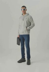 Sweatshirt cinza com capuz e cordão, jeans escuros e ténis pretos. Modelo segurando uma mala de computador preta e usando óculos de sol.