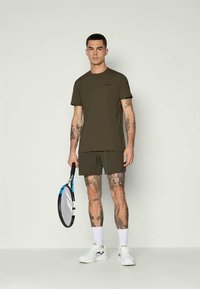 Top sportivo e pantaloni corti verde oliva, miscele di tessuti, design aderente, maniche corte, fronte semplice, abbinati a una racchetta da tennis e calzature bianche.