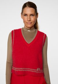 Czerwony, krótki bezrękawnik z dekoltem w serek, wykonany z miękkiego, puszystego materiału. Posiada kontrastujące brązowe wykończenie i linie w pobliżu dolnej krawędzi.