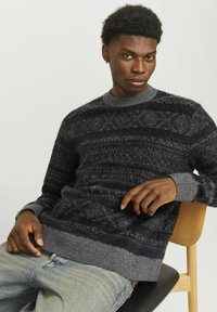 Sweter z dzianiny w ciemnoszarym kolorze z teksturowanymi wzorami, z prążkowanym wykończeniem w okolicy szyi i mankietów. Noszony z jasnoniebieskimi dżinsami, siedząc na drewnianym krześle.