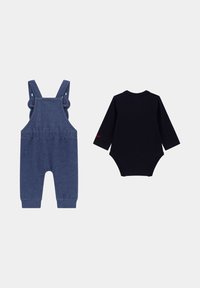 Blaue Jeanslatzhosen mit verstellbaren Trägern und Knopfakzenten, kombiniert mit einem schwarzen Langarm-Bodysuit mit einem runden Ausschnitt.