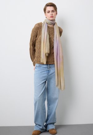 Persona joven con cabello corto que lleva un suéter de punto marrón, jeans anchos de pierna azul claro, zapatos color canela y una bufanda larga con degradado en tonos pastel contra una pared blanca.