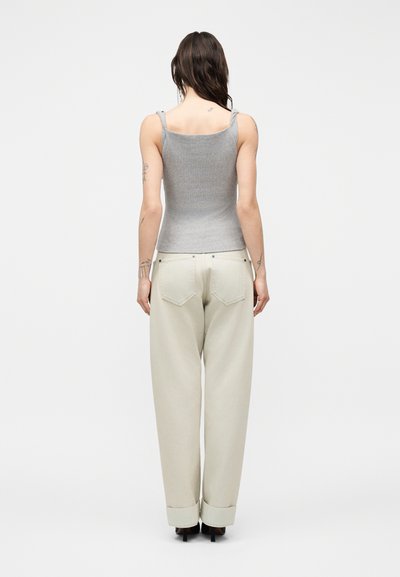 Γυναίκα με μακριά σκούρα μαλλιά, που φορά γκρι μαν sleeveless τοπ και άνετο εκρού παντελόνι, όρθια με την πλάτη προς τα εμάς σε απλό λευκό φόντο.