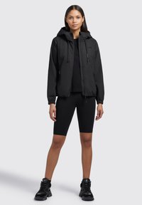 Veste noire légère avec capuche, poignets élastiques et fermeture éclair. Portée sur un haut noir et un short de cyclisme noir, accompagnée de chaussures noires.