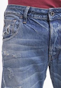 Jeans de mezclilla azul desgastados llevados con una camisa granate metida por dentro, mostrando detalles del frente de la cintura y los bolsillos.