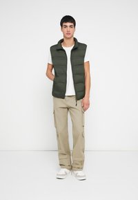 Olivengrønn vattert vest med høy krage, båret over en hvit t-skjorte, kombinert med beige cargobukser og hvite joggesko.