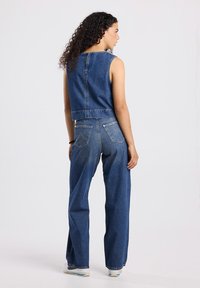 Woman with curly hair wearing sleeveless denim top and wide-leg blue jeans, standing sideways against a plain light background.