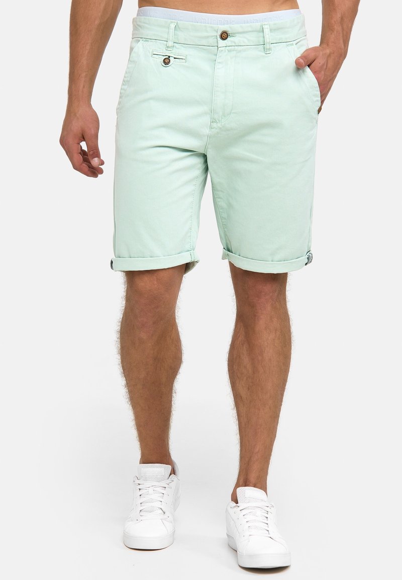 Homme portant un short vert clair roulé, les mains dans les poches, des baskets blanches, debout sur un fond uni clair.
