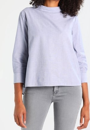 Femme portant un chemisier bleu clair à manches longues avec un col montant et un jean ajusté gris clair, debout devant un fond blanc.
