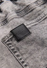 Tela de mezclilla gris con una etiqueta rectangular negra cosida que dice "ombre" cerca de un remache metálico en la esquina de un bolsillo.